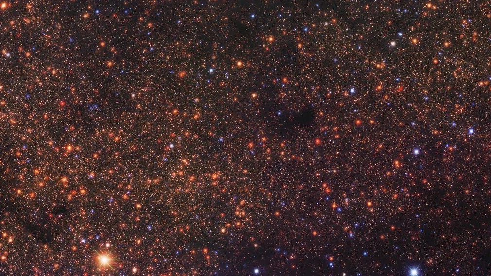 A wide-angle image of the universe showing thousands of stars in reddish, orange, and bright blue points of light. 