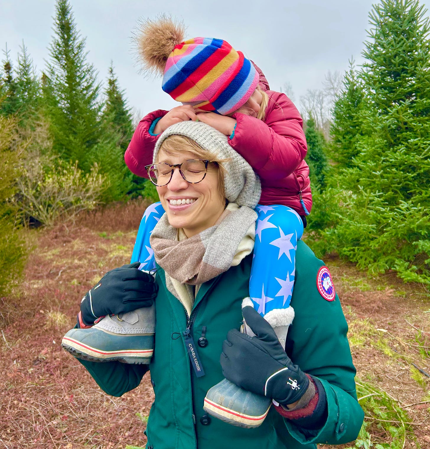 Susan with her daughter on her shoulders at a Christmas tree farm with a gray sky and green fir trees. Susan is smiling, her daughter's face is hidden to the side.