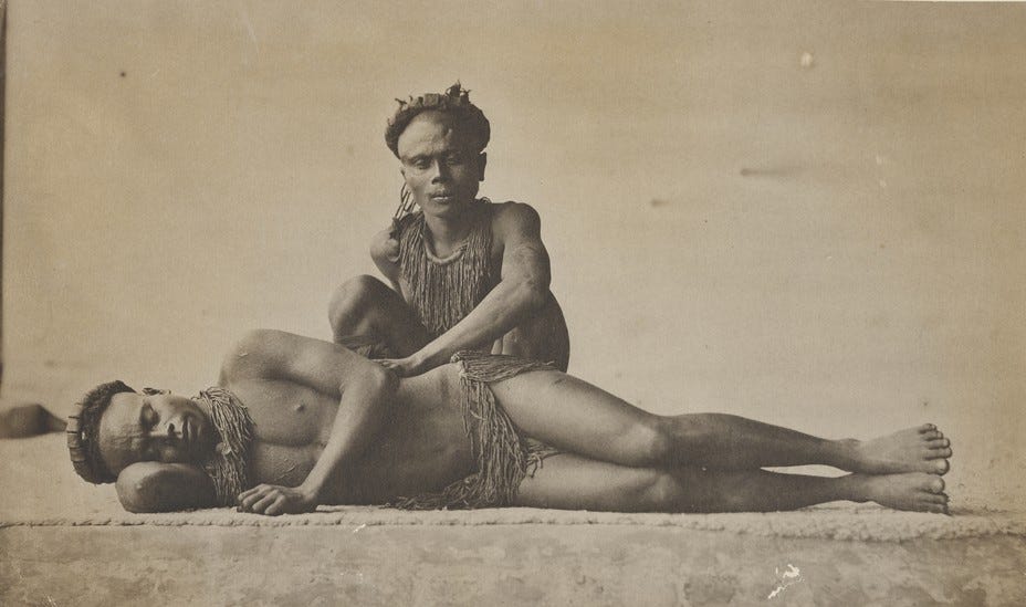 Two Andamanese men shown, one lying on the ground and the other with his hand on his side.