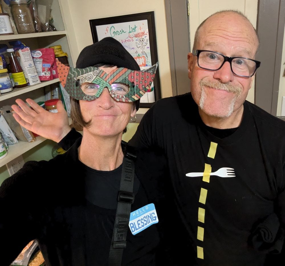 Any and me in the kitchen of a house. I am wearing all black with a mask over my eyes and a sticky nametag that reads "Blessing." Andy is wearing all black with a yellow line down the front of his shirt. There is a fork taped in the middle of the shirt.