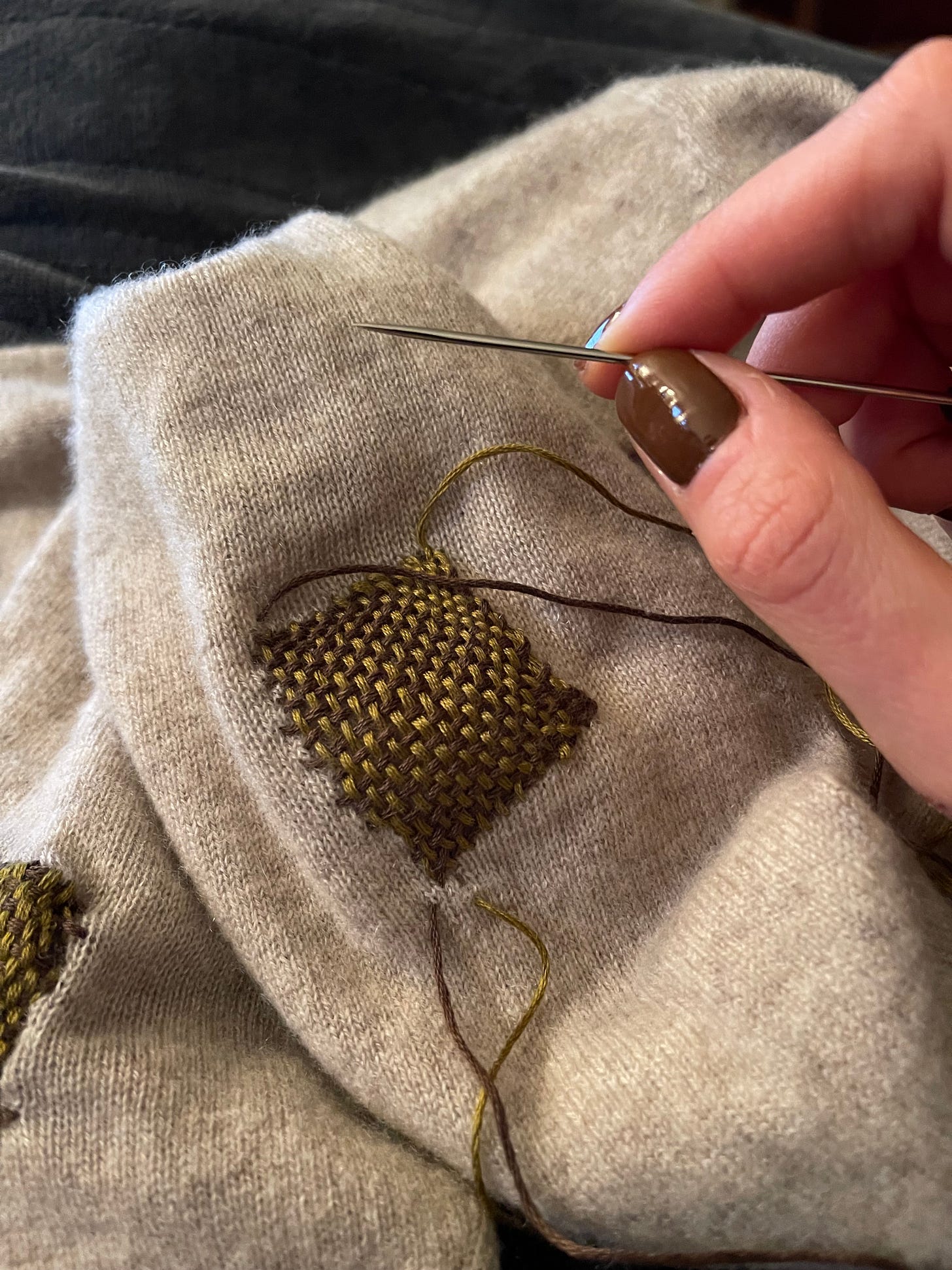 Hand holding darning needle with beige patched sweater in the fore