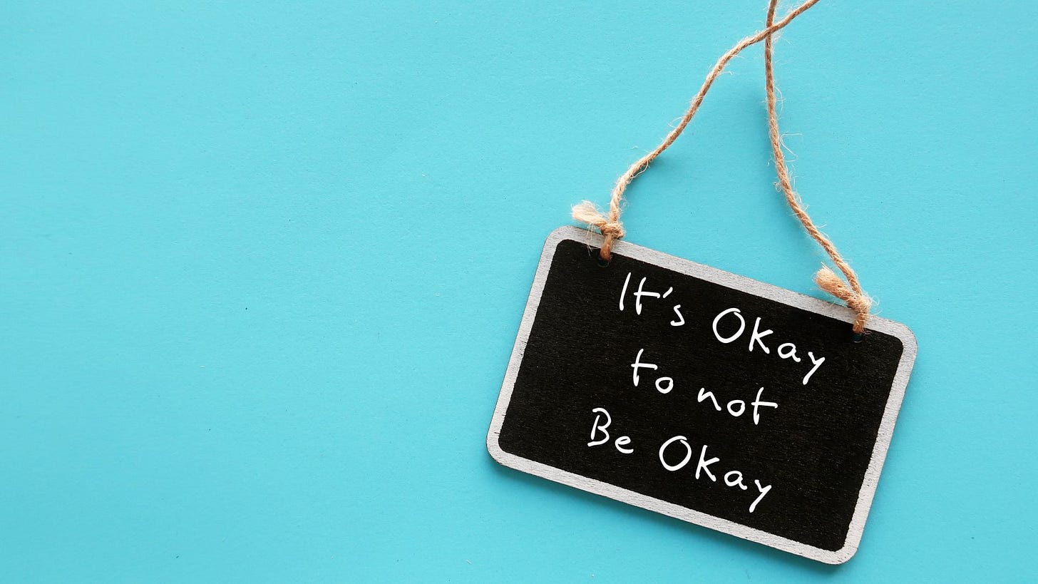 Sky blue solid background, small blackboard with white border hanging on twine with writing that says “It’s Okay to not Be Okay”