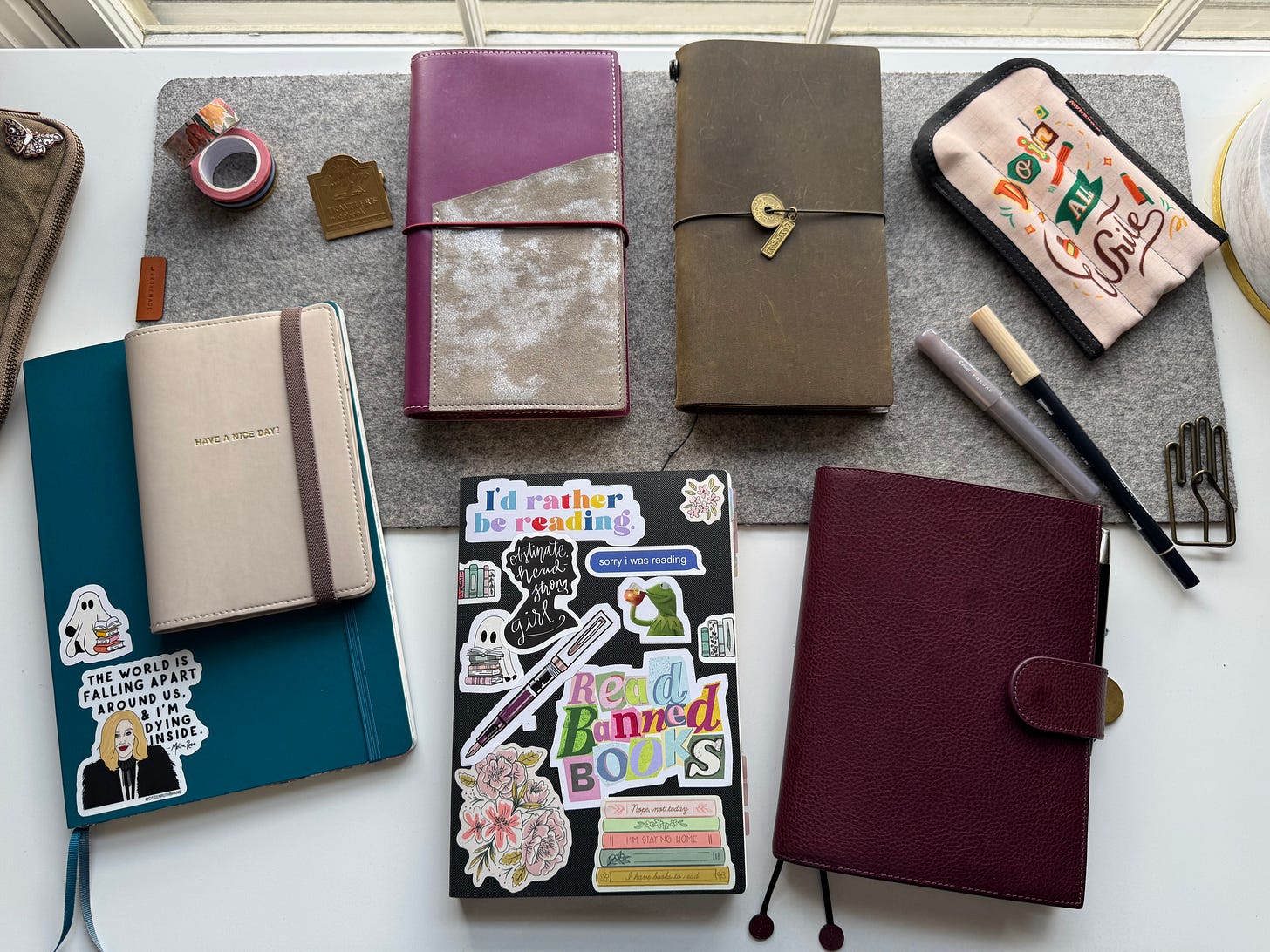 Six notebooks shown on a white desk, with a grey felt desk mat. From Left to Right: B5 Leuchtturm 1917 Soft cover notebook (Color: Pacific Green), A6 Notebook with Hobonichi “Have a Nice Day!” Cover, Standard size notebook in BujoPlanner cover, Standard size traveler’s notebook (Color: Olive green), A5 Sterling Ink Grid Notebook with reading related stickers on the cover, B6 notebooks with Gillio Firenze Appunto Cover (Color: Epoch Purple). 