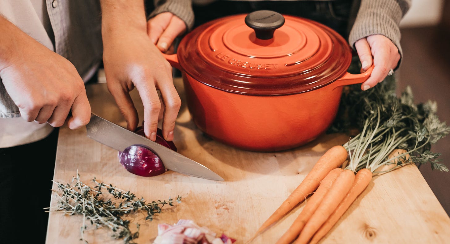 Orange enamel pot with lid on cutting board with two hands on handles, while two more hands slice a red onion. 