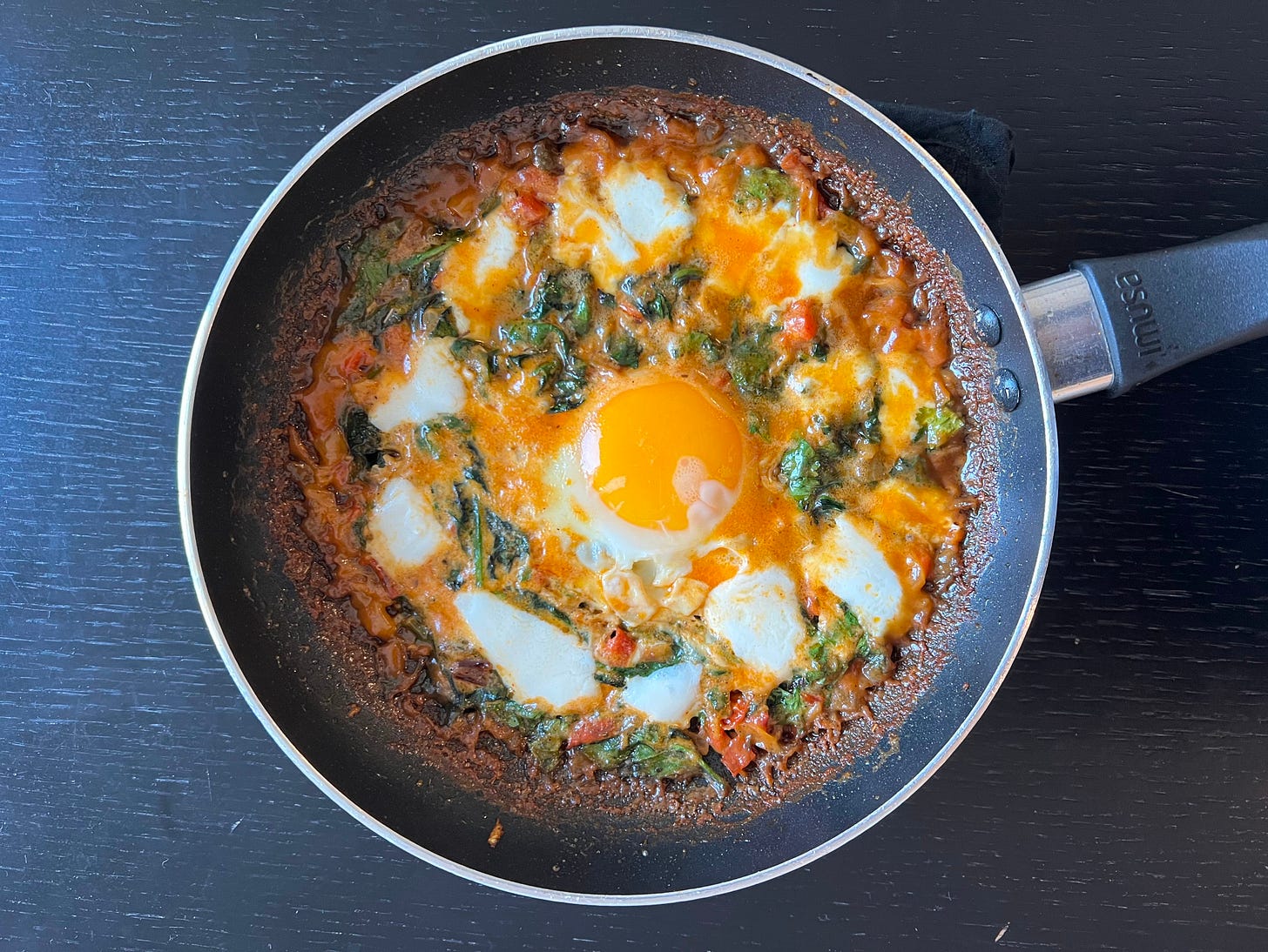 A homemade shakshuka with a rich, tomato-based sauce, fresh spinach, and melted cheese, crowned with a perfectly cooked egg in the center. The colors of the dish pop against the dark surface, highlighting the contrast between the golden yolk, red tomatoes, and green spinach. Another Foodie Disorder experiment by Liat Portal, crafting a shakshuka variation that brings together fresh ingredients and bold flavors.