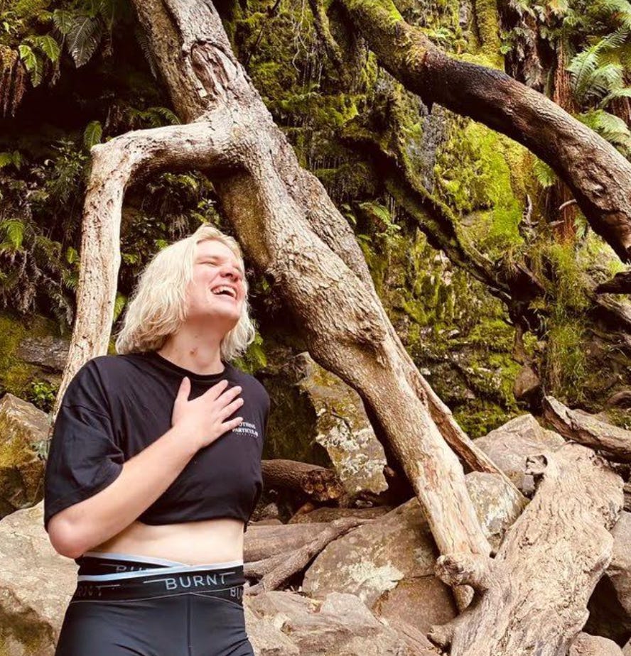  A blonde non-binary person holding their hand to their chest and laughing in front of a waterfall
