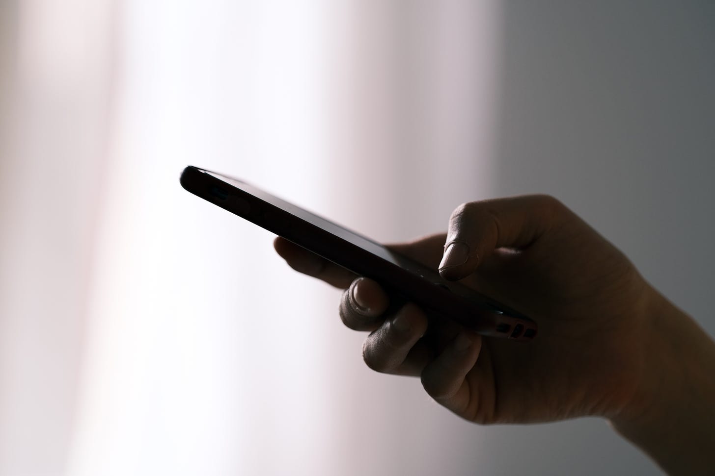 The silhouette of a person's hand texting on a black phone. The background is a white curtain with light coming through.