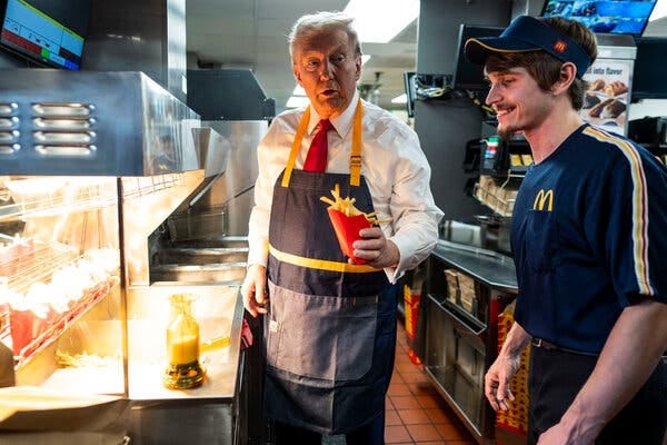 Donald Trump, in a white shirt and red tie with a dark apron covering his outfit, holds up a red carton of fries while looking at a McDonald’s employee who is smiling.