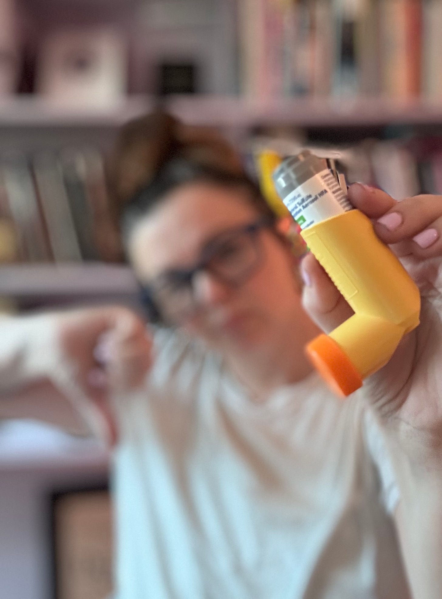 Amber holds up a yellow inhaler which is in the foreground of the image. In the background, Amber is out of focus, but you can see she is doing a "thumbs down" gesture. 