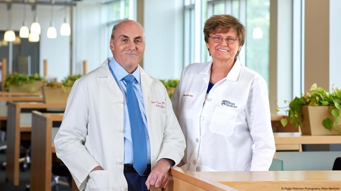 Nobel Prize winners Drew Weissman (left) and Katalin Kariko stand next to each other in an office. Both are wearing white lab coats and smiling at the camera.