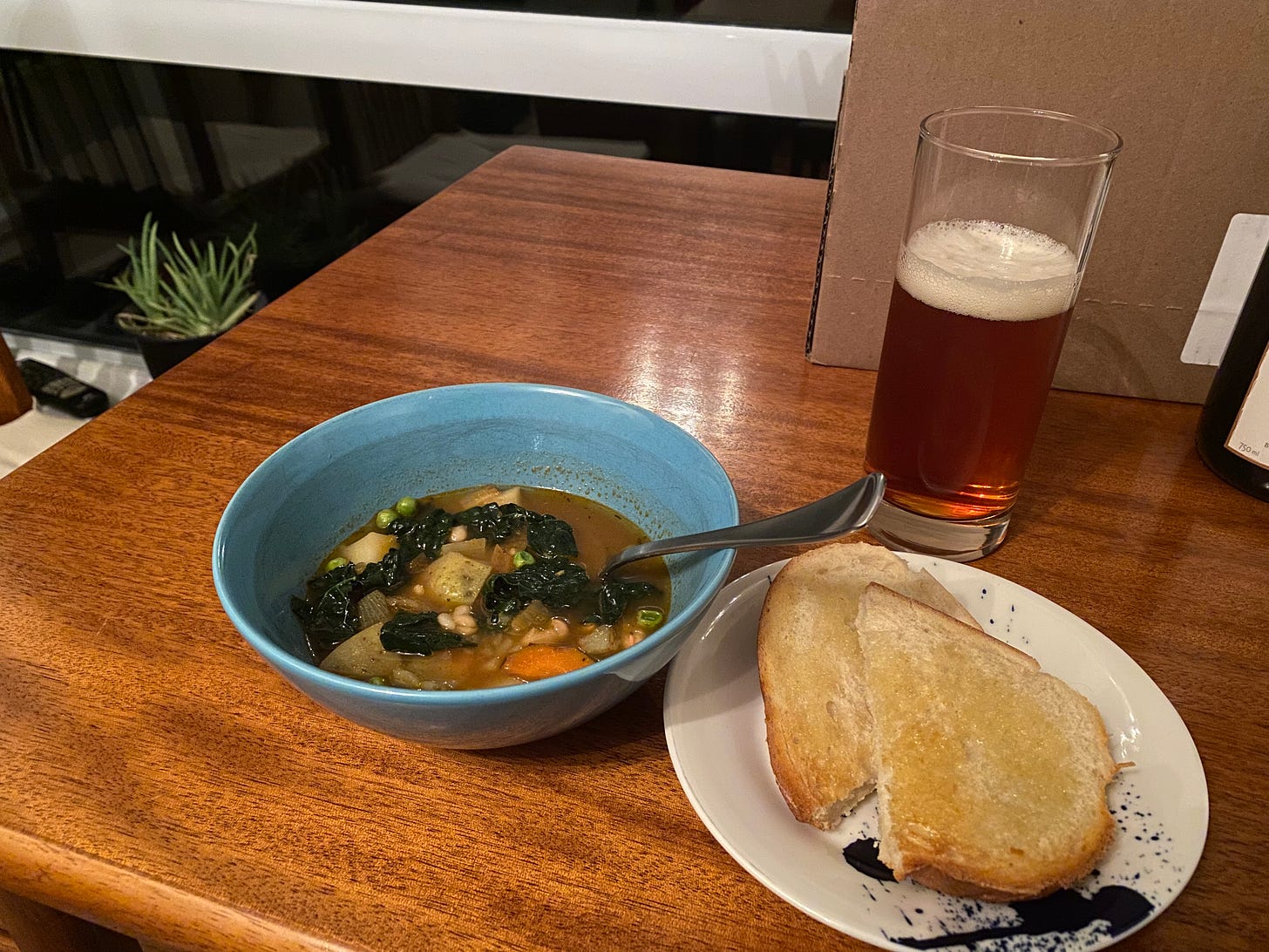 A blue bowl of chunky vegetable soup next to a small plate with buttered toast, and a tall glass half-full of beer behind them.