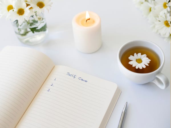 Journal opened to a page titled Self-Care, next to a lit white candle, a cup of tea with a flower in it, and a jar holding white daisies