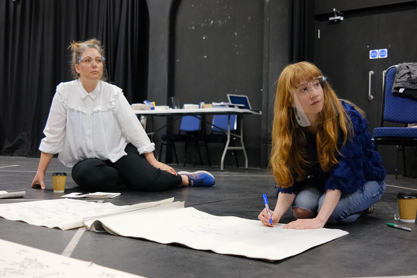 Two women in transparent masks sit in a rehearsal room on the floor beside big notepads of paper full of writing