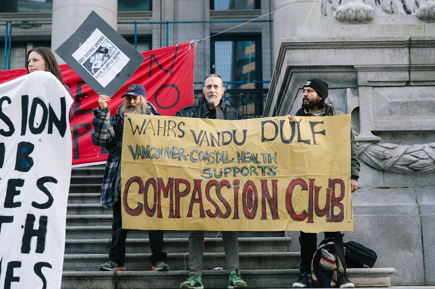 Three people on stairs at the Vancouver Art Gallery hold banners, one saying “WAHRS VANDU DULF Vancouver Coastal Health supports Compassion Club,” another saying “No more dying” and “safe supply now”