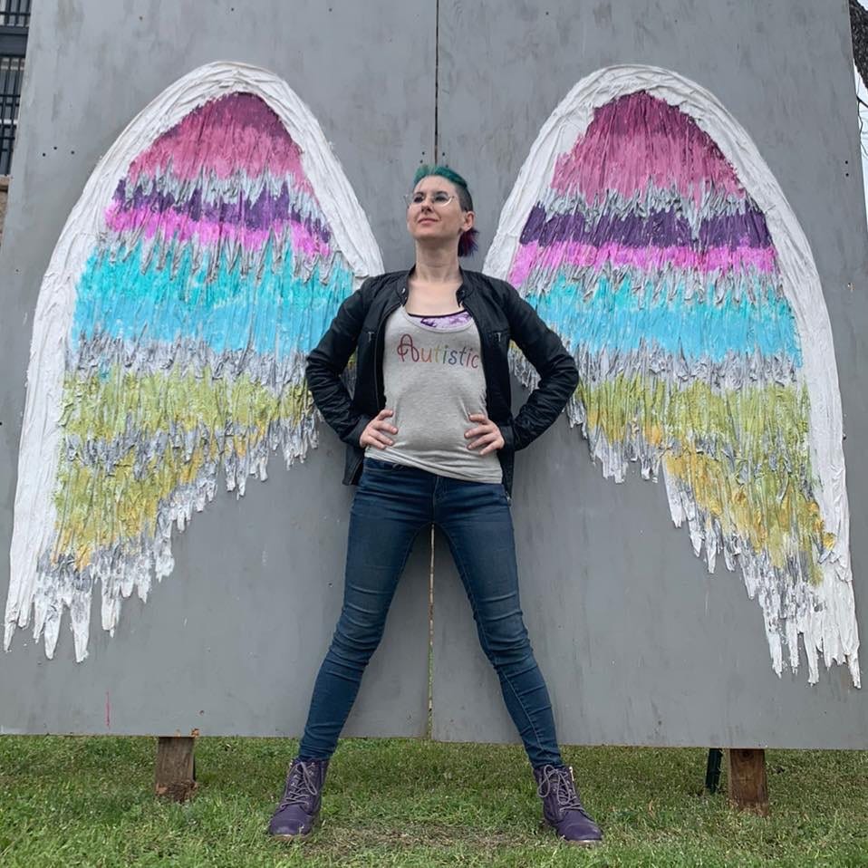 Photo of Lyric, in purple boots, jeans, a grey tank top that says “Autistic” in a rainbow font on the front, and a black leather jacket, posing in a power-pose in front of some painted wings.