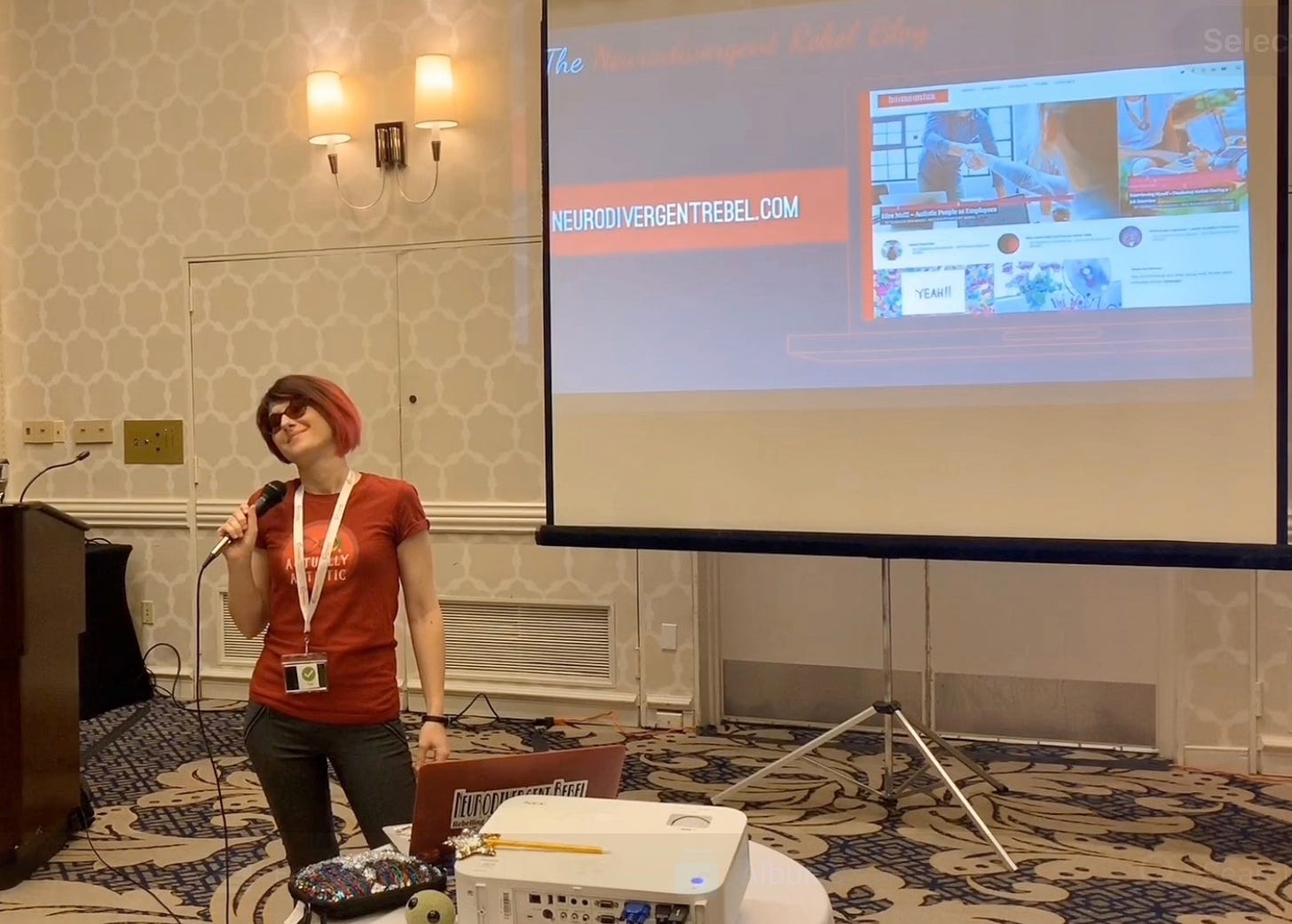 Lyric Rivera, in a red ActuallyAutistic shirt, stands on the floor in front of a projector, holding a microphone, smirking, and tilting their head. 