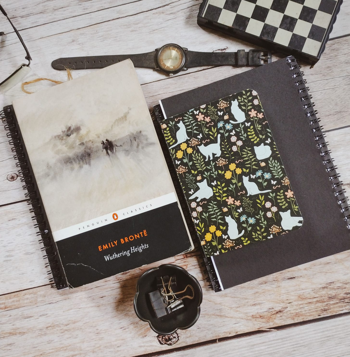 Copy of Emily Brontë's Wuthering Heights, on a wooden tabletop. Also on the tabletop are two notebooks, an inkstand, binder clips, an old watch and a chessboard