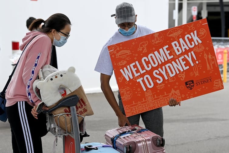 A man wearing a mask holds a sign saying 'Welcome back to Sydney'. A woman in a mask wheels a trolly full of suitcases.