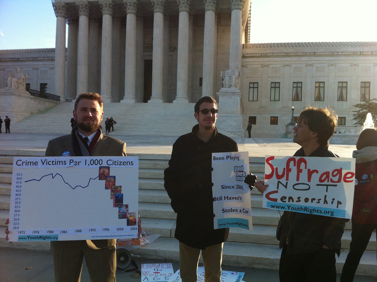 Three men standing in front of the U.S. Supreme Court building holding signs. The middle one states: "“Been playing Grand Theft Auto since 2004. Still haven’t stolen a car.”