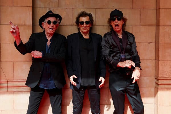 Keith Richards, Ronnie Wood and Mick Jagger, all in sunglasses, stand next to a wall in shadows.