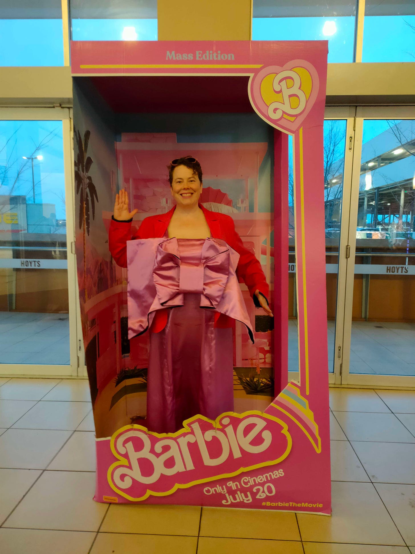 The author, a white woman with short dark hair, stands in a box designed to look like Barbie doll packaging. She wears a dark pink blazer-style jacket and a mid-pink shiny long gown with a low square neckline. The dress sports a truly gigantic 3D bow that has a ton of interfacing to make it stiff.