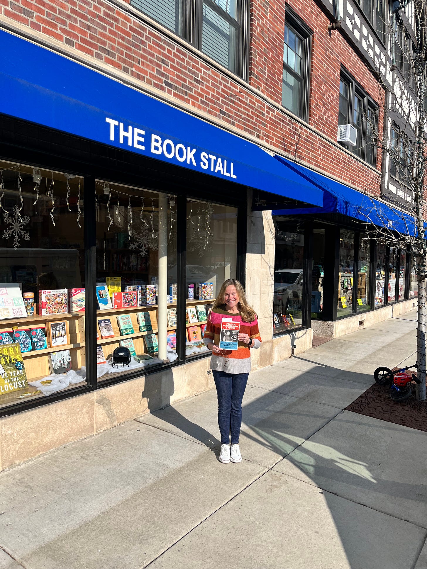Memoir Coach Christine Wolf stands outside of independent bookstore The Book Stall in Winnetka, Illinois, holding her debut book, Politics, Partnerships, & Power: The Lives of Ralph E. and Marguerite Stitt Church. She's wearing a striped sweater and denim jeans. The bookstore has a red brick facade and a blue awning with white letters, and the store windows are filled with books and twinkle lights left over from the recent holidays. 