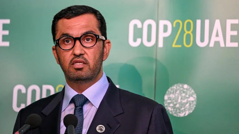 Image of Sultan al Jaber in a suit and tie with glasses in front of the COP28 green branded sign.