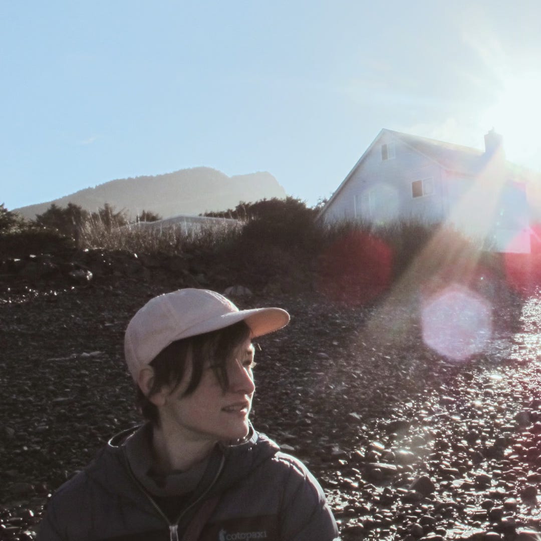 A photograph of Sarah, looking to the right of the frame and wearing a pink baseball cap over short hair. She is standing on a rocky short at the beach. A house, some plant matter, and a mountain can all be seen in the distant background.