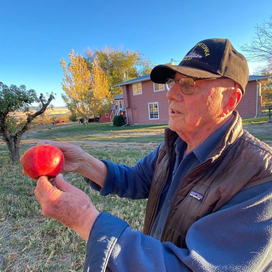 Man wearing a hat holding up a Rome Beauty red apple on a crisp autumn day, clear blue sky, golden trees and home in the background.
