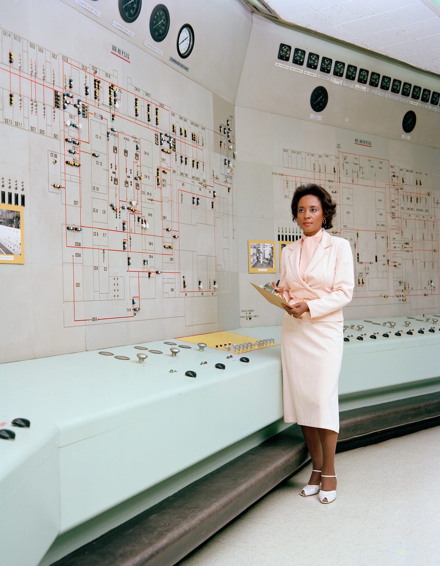 Annie Easley wearing a pink skirt suit and white low heels inside a computer room with clocks lining the top of the room.
