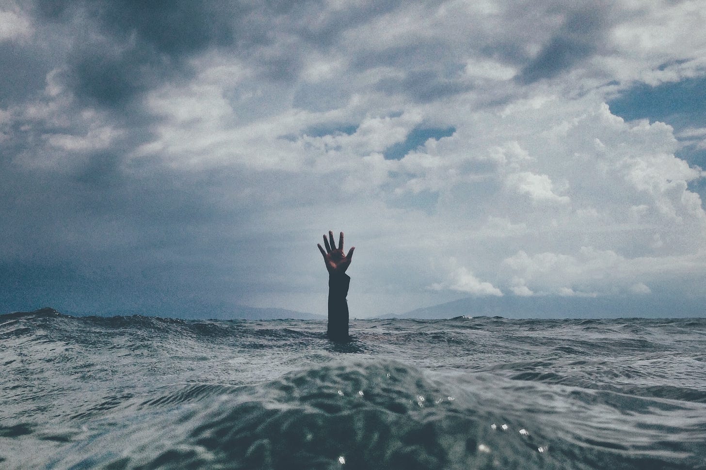 A person's hand sticks out of a churning sea. The hand and arm are visible up to the elbow. Above is a cloudy, dark sky.