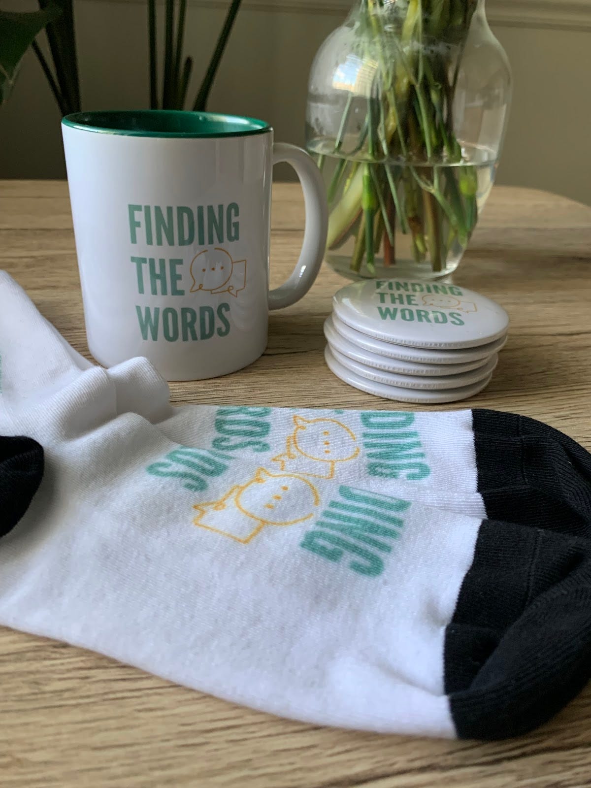 A white mug with a green interior, a stack of six circular magnets and a pair of socks on a wooden table. They are all white with “Finding the Words” written in green along with an orange drawing of text message bubbles–it is the logo for Finding the Words. There is a vase and a plant in the background.