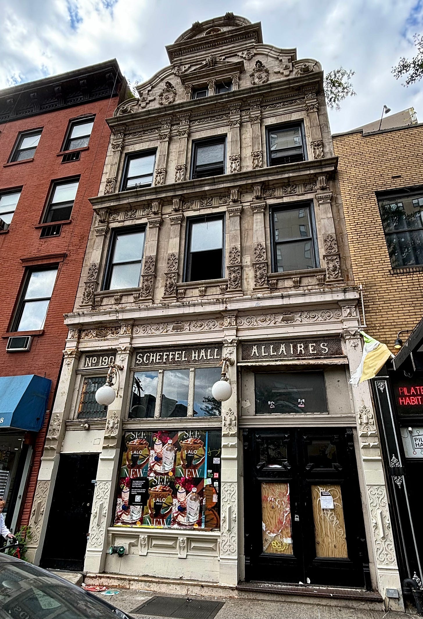 An ornately decorated gray stone building in a state of disrepair. Over the door is a decorate colored window and the marking No 190. Over the main storefront entrance it is labeled Scheffel Hall/Allaires. There are adds plastered over the front window.