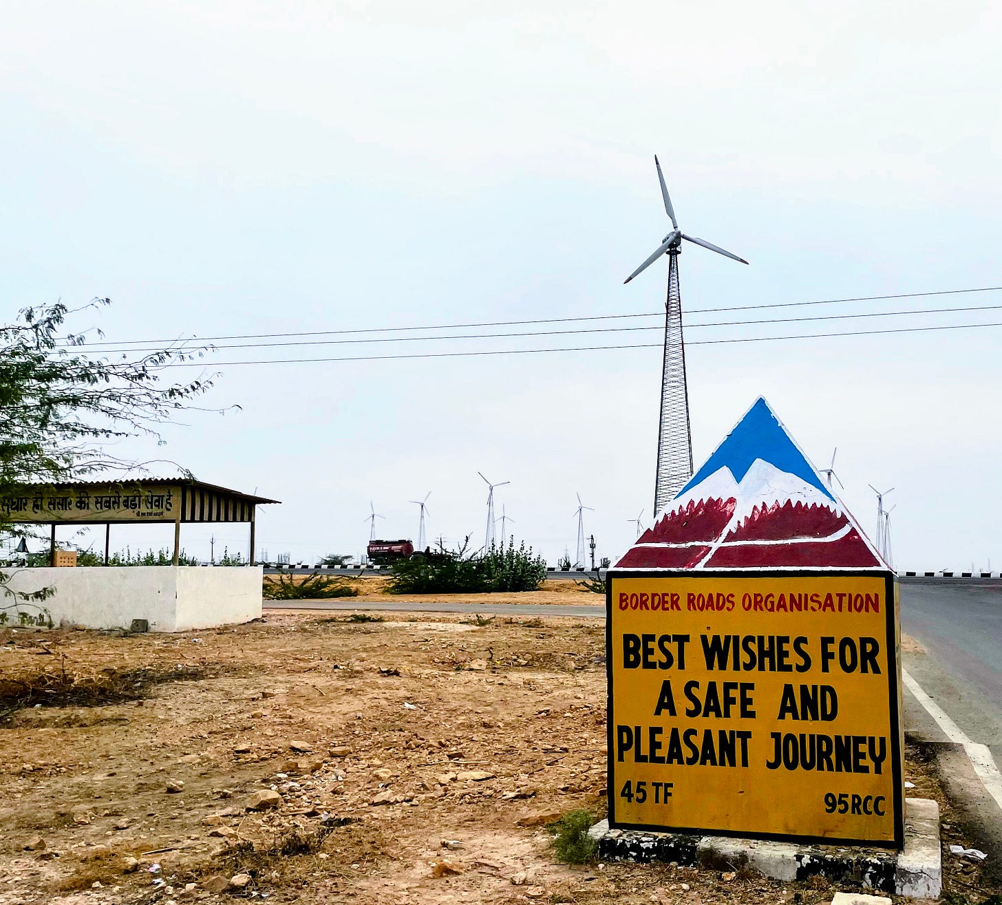 A sign by a road in India wishes drivers "Best wishest for a safe and pleasant journey." Behind the sign are many large wind turbines and electrical wires.