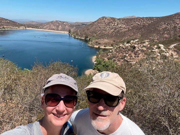 Selfie of a man and woman wearing ball caps and sunglasses smiling in front of a lake in Southern California