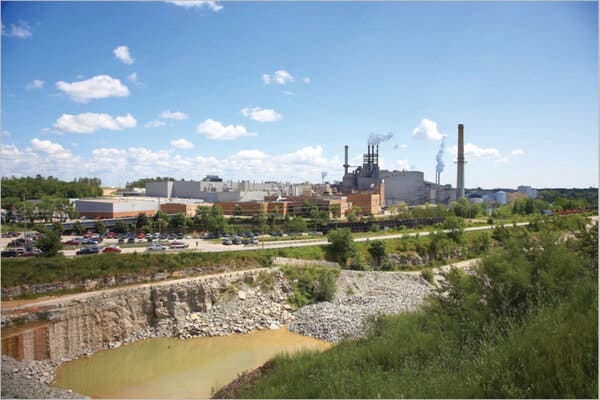 A paper mill plant with chimneys in the background and a large stone pit filled with water in the foreground.