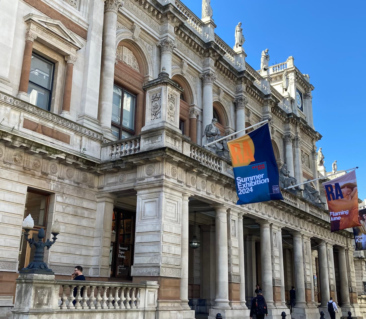 Burlington House against a blue sky. the building is highly decorated architecture with columns, deep friezes, and statues. Flags hang above the entrance announcing the Summer Exhibition.