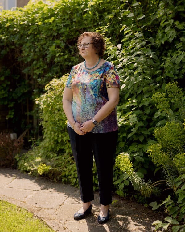 Hilary Cass standing near a bush with her hands clasped before her on a sunny day. She wears a colorful shirt and black slacks.