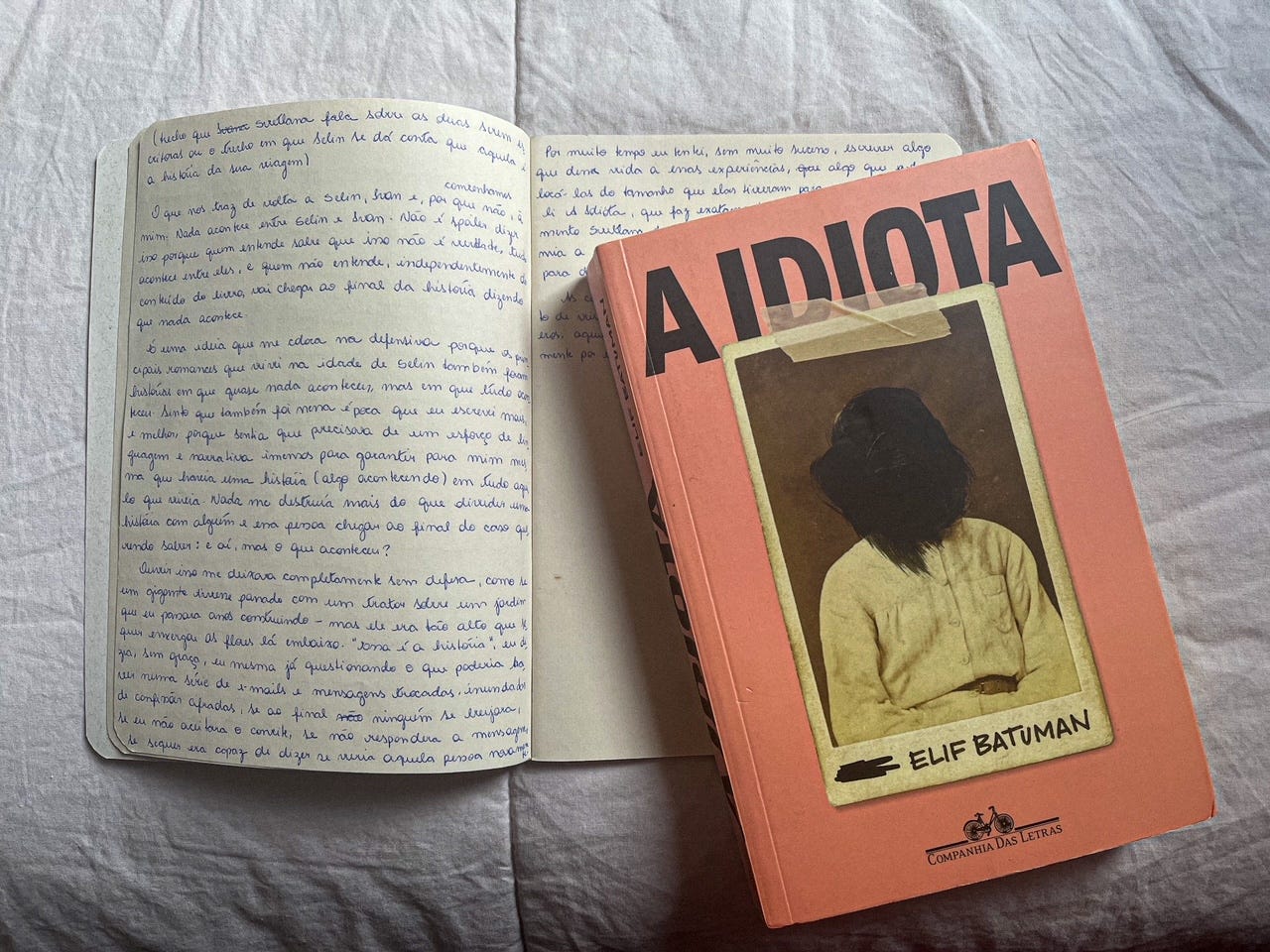 Caderno aberto sobre fundo lilás, com o primeiro rascunho desse texto, ao lado do livro fechado