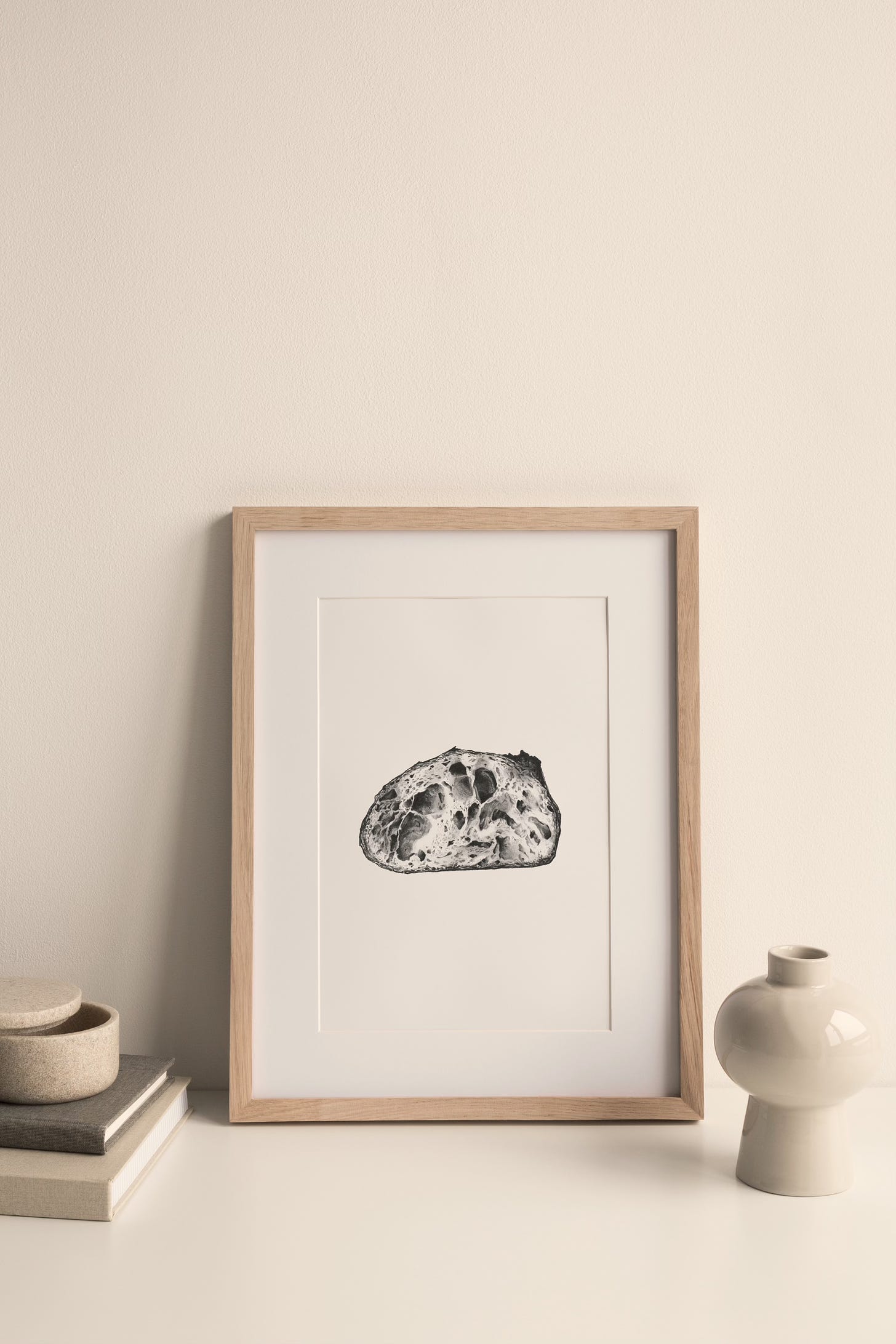 A sourdough pencil drawing in a frame, sitting on a surface with some books to the left and a vase to the right