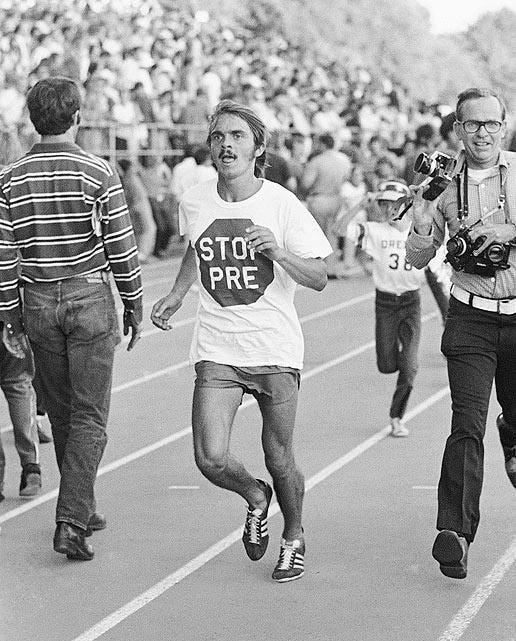 Black-and-white photo of legendary American distance runner Steve Prefontaine during a victory lap on a track. He wears a white t-shirt with a mock stop sign that reads “STOP PRE,” symbolizing his dominance in racing. Prefontaine is mid-stride, looking determined, with a mustache and shaggy hair. A crowd fills the bleachers in the background, while a cameraman and other figures walk alongside him, capturing the moment. The scene is iconic of 1970s track and field culture and Prefontaine’s larger-than-life persona.