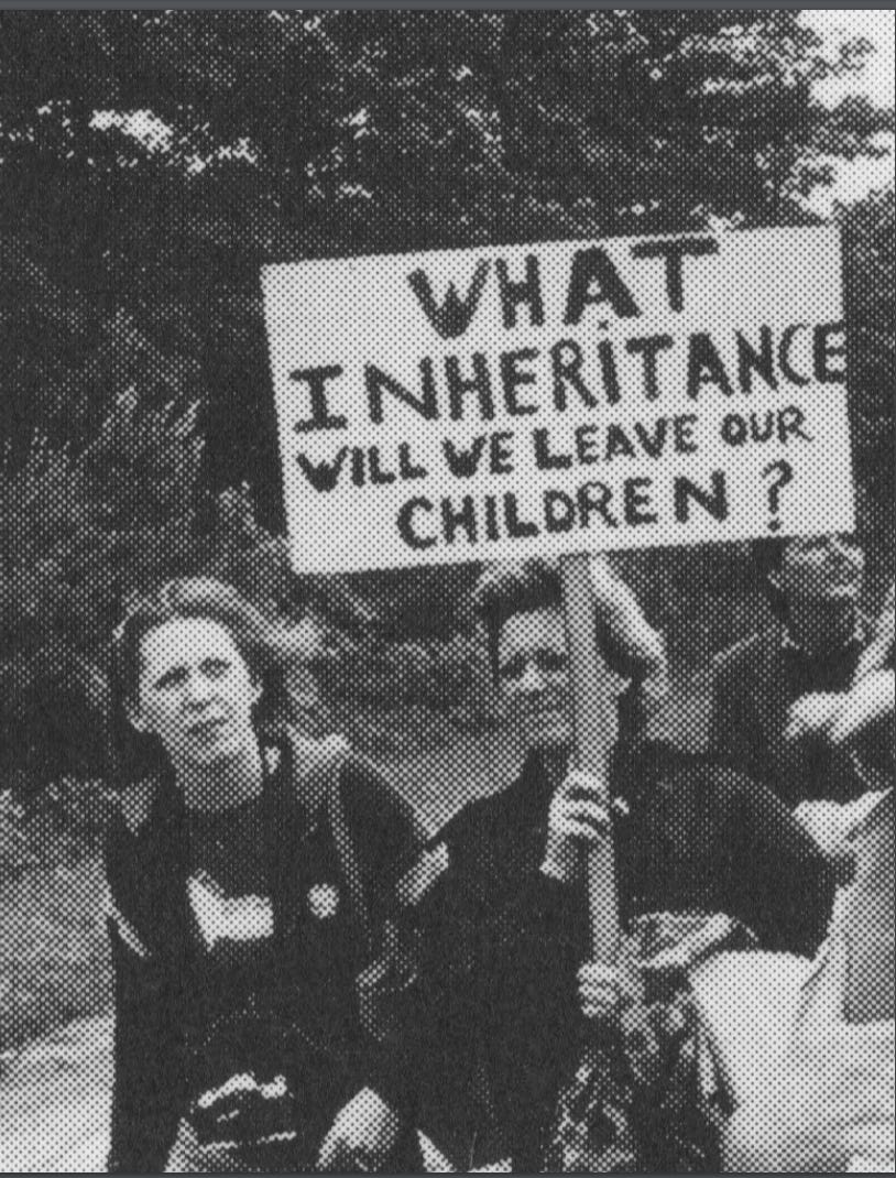 Black and white photo of a white women holding up a sign that reads "What inheritance will we leave our children?" She is walking next to another white women. It looks like it is windy. They are walking by some tall trees.