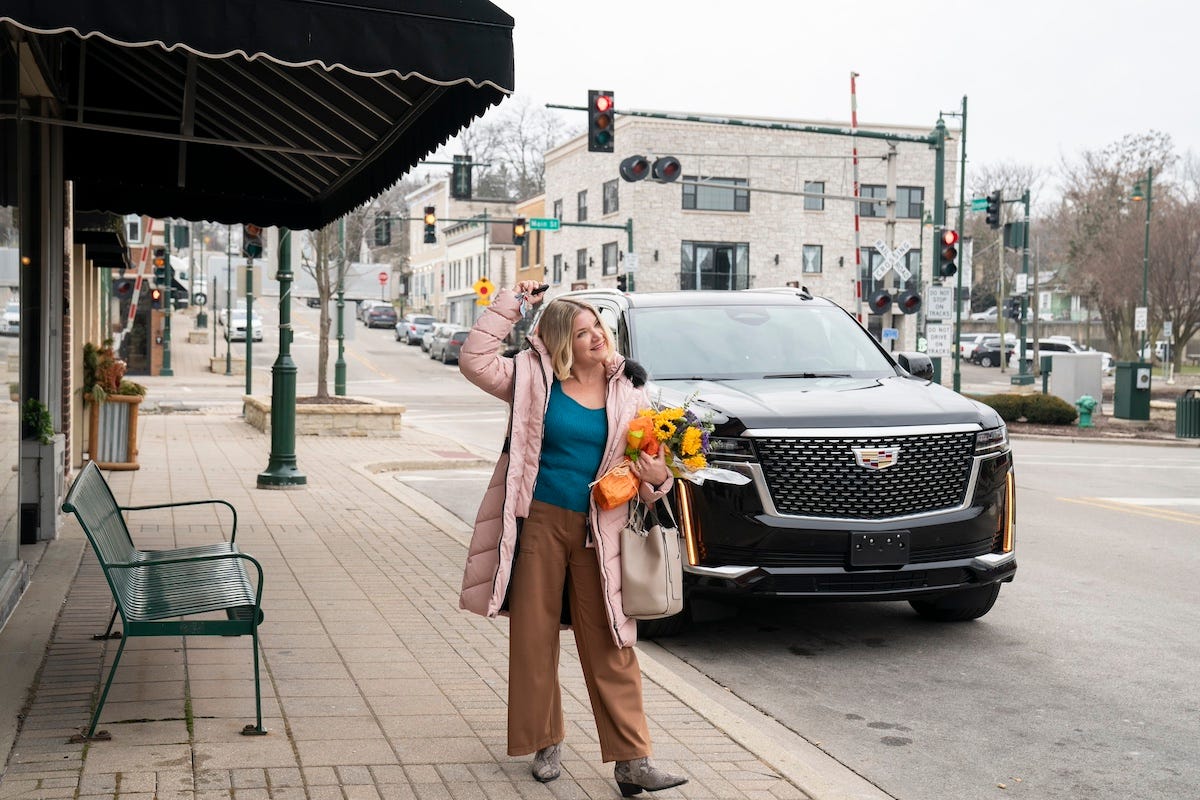 Mary Catherine Garrison shown on a street in a pink jacket waving in front of a Cadillac Escalade