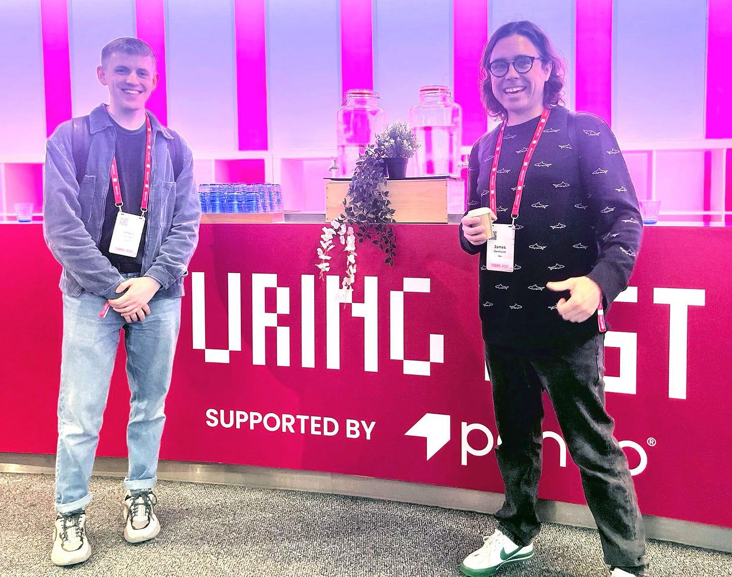 James and Lewis standing in front of a brightly-lit Turing Fest sign, smiling.