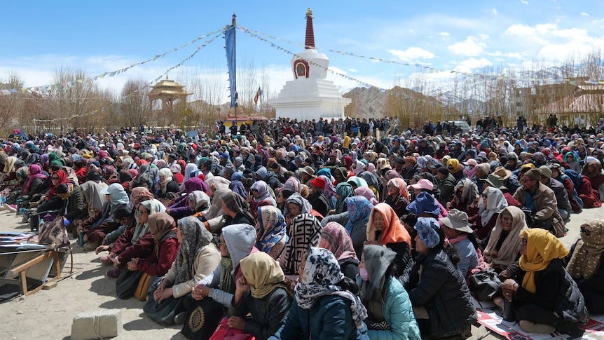 Thousands of people in shawls sit on the ground near a monument under blue sky