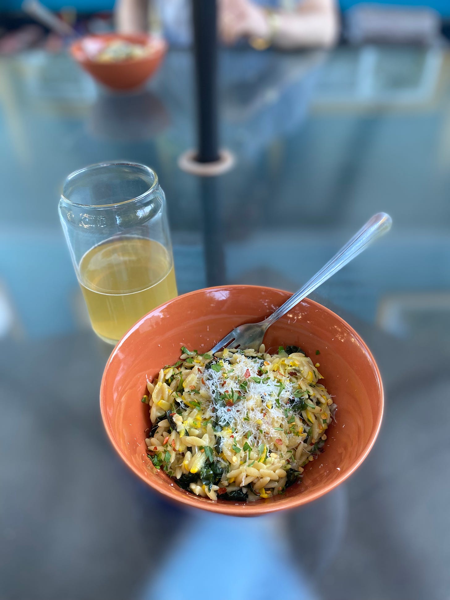 An orange bowl of the orzo described above, dusted with parmesan and chili flakes. A fork sticks out at the back of the bowl, and next to it is a pint glass of pale ale.