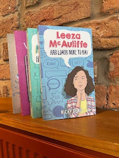 Four books, standing on a wooden bookshelf. All four spines can be seen, but only the front cover of the book at the front of the row, is visible. That front cover is the new book, Leeza McAuliffe Has Loads More To Say by Nicky Bond. The other books are Leeza McAUliffe Has Loads More To Say, then Assembling the Wingpeople, and then Carry the Beautiful at the back. All books are by Nicky Bond and the name can be seen on the spines.