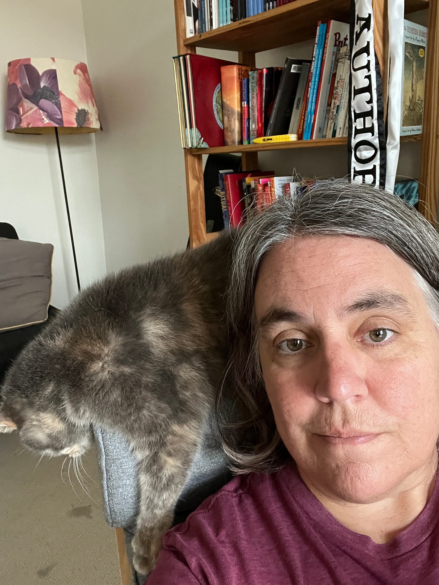 Selfie of Robyn in her writing chair, with a gray cat balanced behind her on the chair, hanging off and looking bored