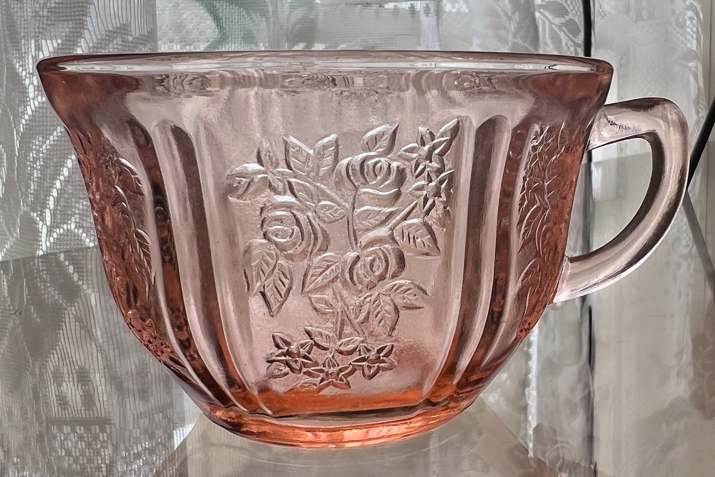 A pink depression era coffee cup sitting on a glass shelf with lace curtains in the background