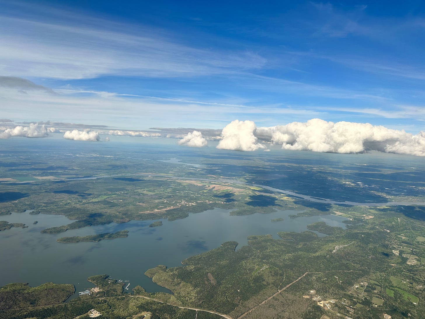 View from airplane overlooking Little Rock Arkansas region at cloud level. Water and green below, blue sky above, flat-bottomed white clouds splitting the view horizontally in the middle. 